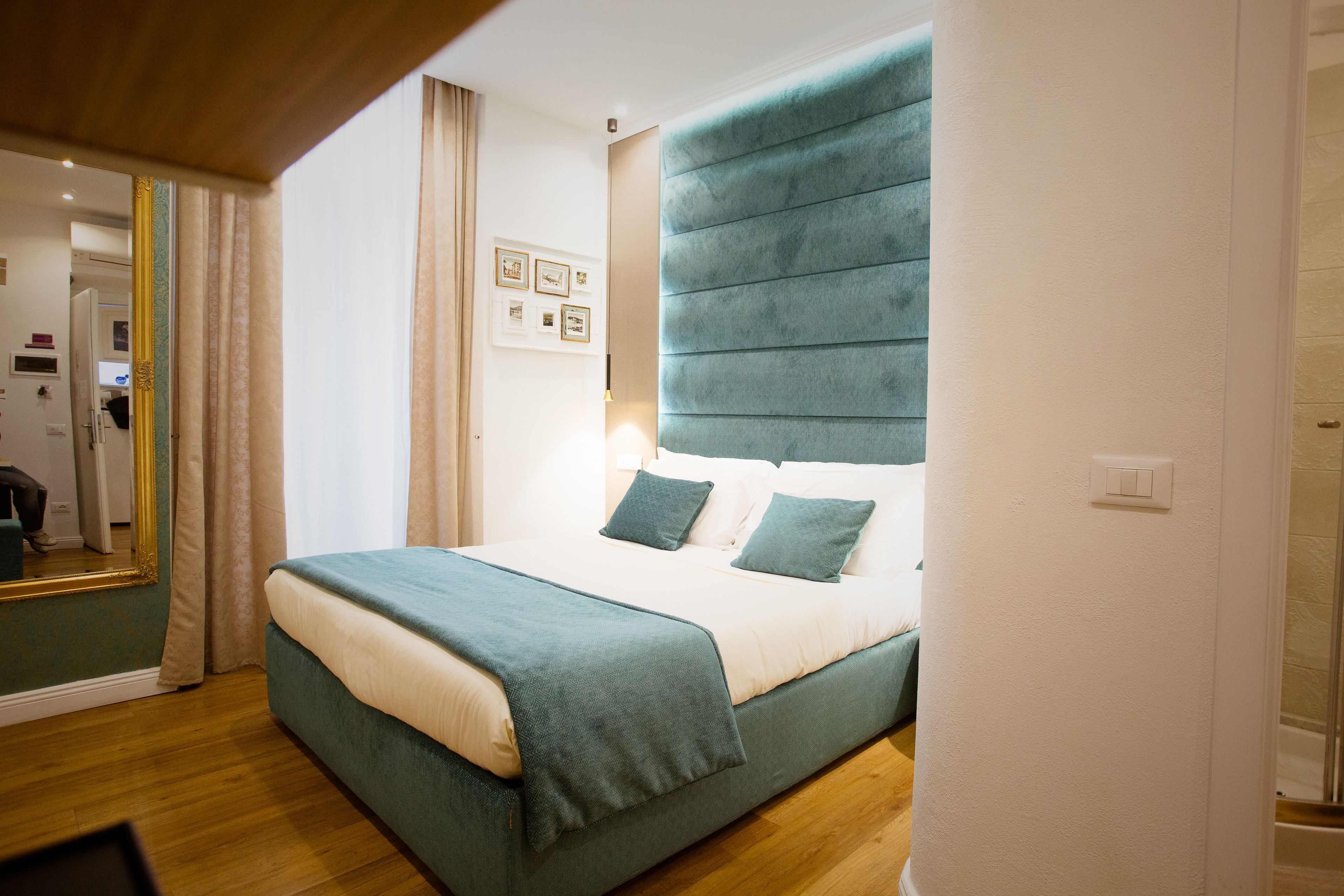 A neatly made bed with a teal headboard and pillows, a wooden floor, and a mirror on the left.