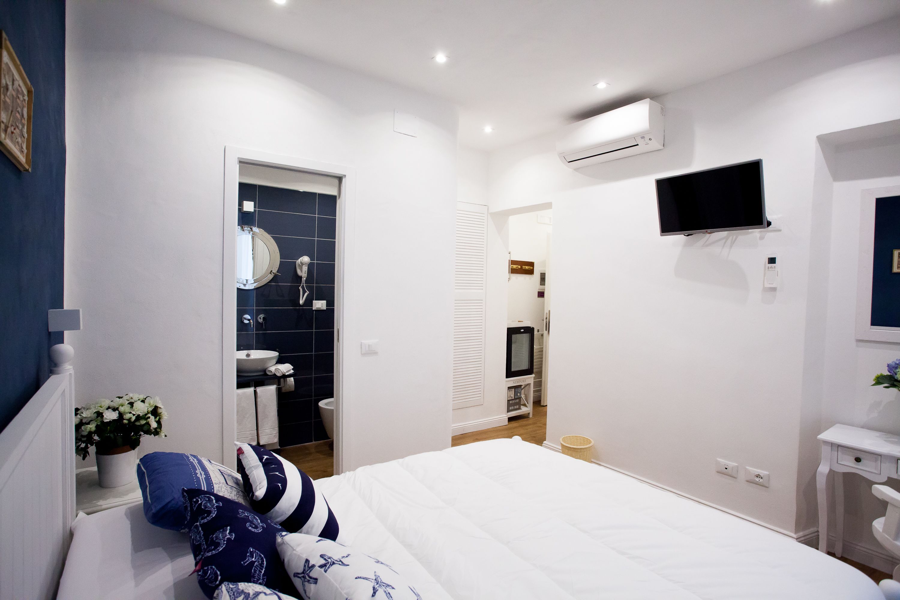 Une chambre à coucher avec un lit, une table de chevet ornée d’une plante, un téléviseur fixé au mur, et une salle de bain visible à travers une porte ouverte.