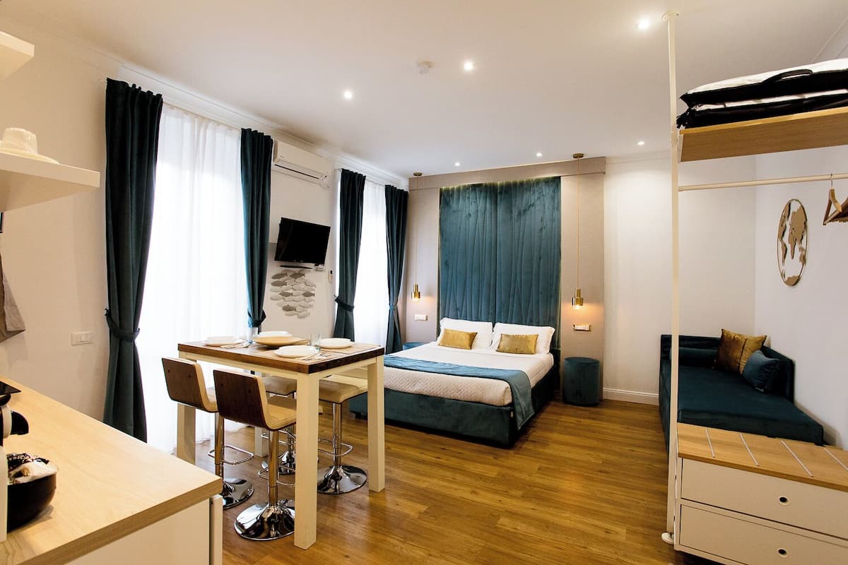 Ein modernes Schlafzimmer mit einem Bett, einem Schreibtisch und einem Sessel. Ein Fernseher ist an der Wand montiert und es gibt einen kleinen Tisch mit Stühlen.