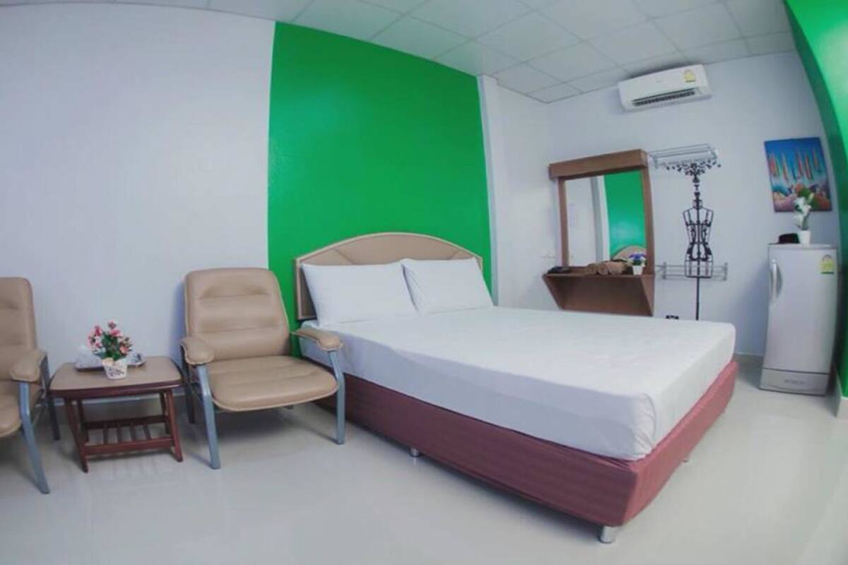 A hotel room with a bed, two chairs, a small table with a vase, a mirror, a wardrobe, and a refrigerator.