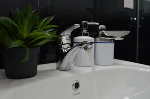 Un lavabo de salle de bain avec un robinet ouvert, une plante en pot et un récipient blanc.