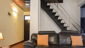 A modern living room with a black leather sofa, two orange cushions, a glass coffee table, and a staircase with metal railings. - Panoramic Holiday Apartment Seagull Complex - Nuwara Eliya (Nuwara Eliya)