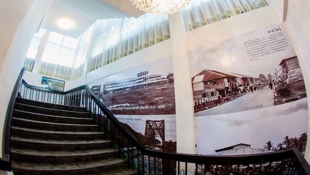 Uma escadaria com um candelabro, um cartaz grande com fotografias históricas e vista para um edifício ao fundo.