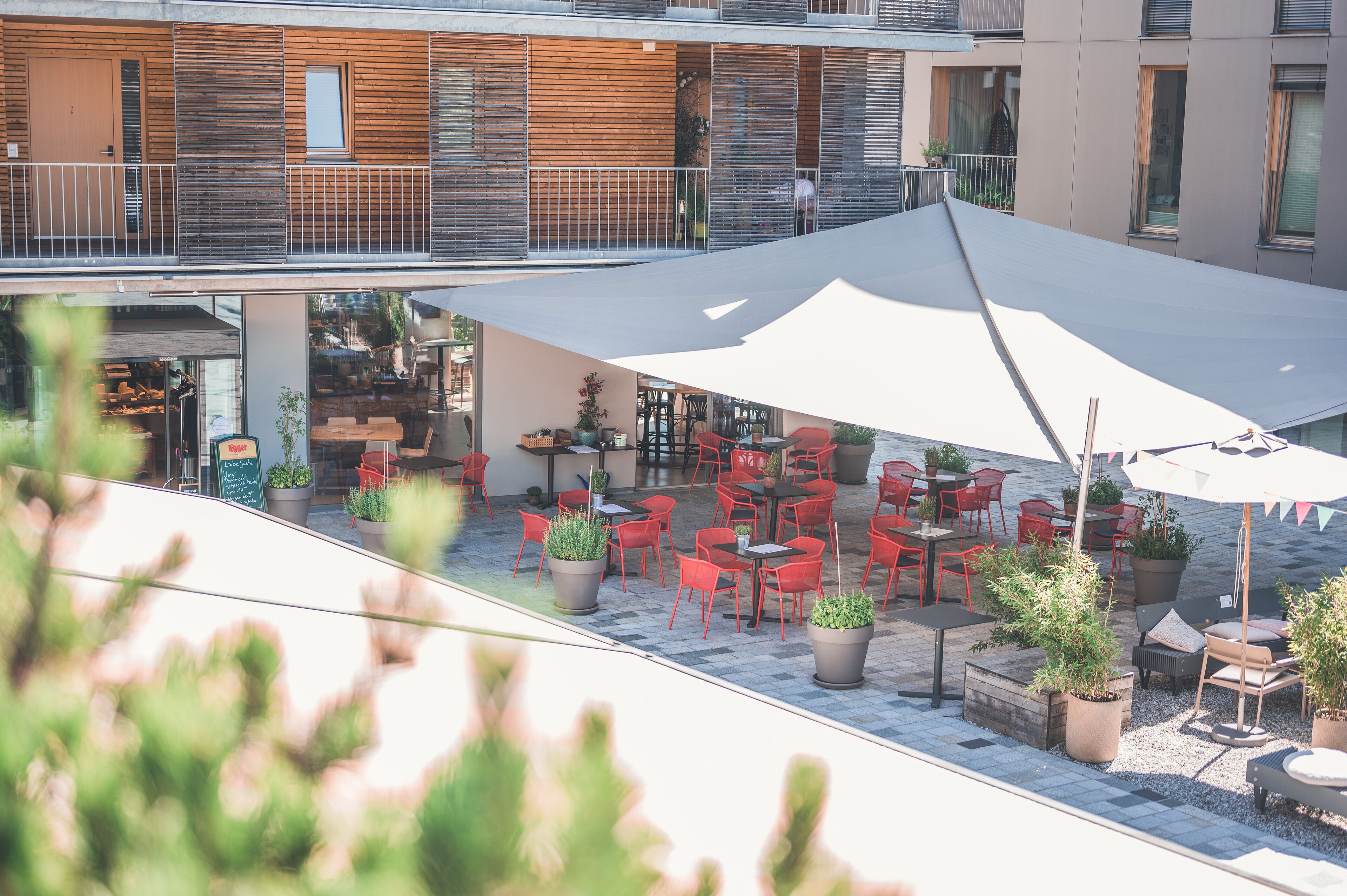 Un espace extérieur aménagé avec des chaises et des tables rouges, entouré de plantes en pot et surmonté d’un grand parasol.