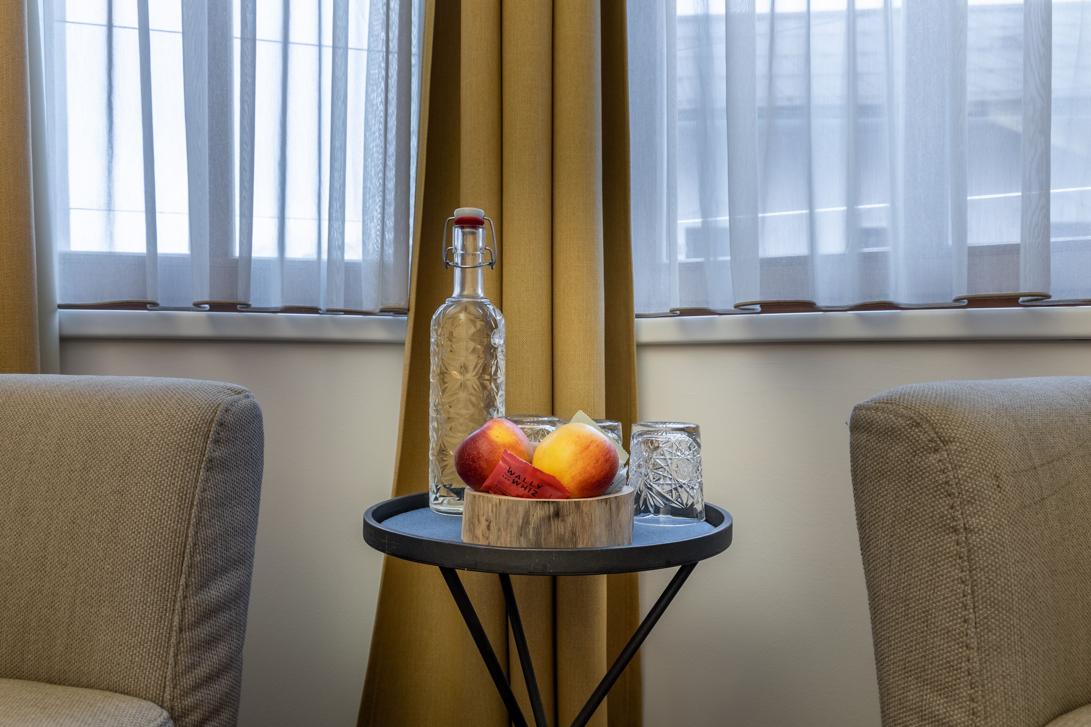 Habitació amb una taula petita que sostené una ampolla, dues copes i un bloc de fusta amb fruites.