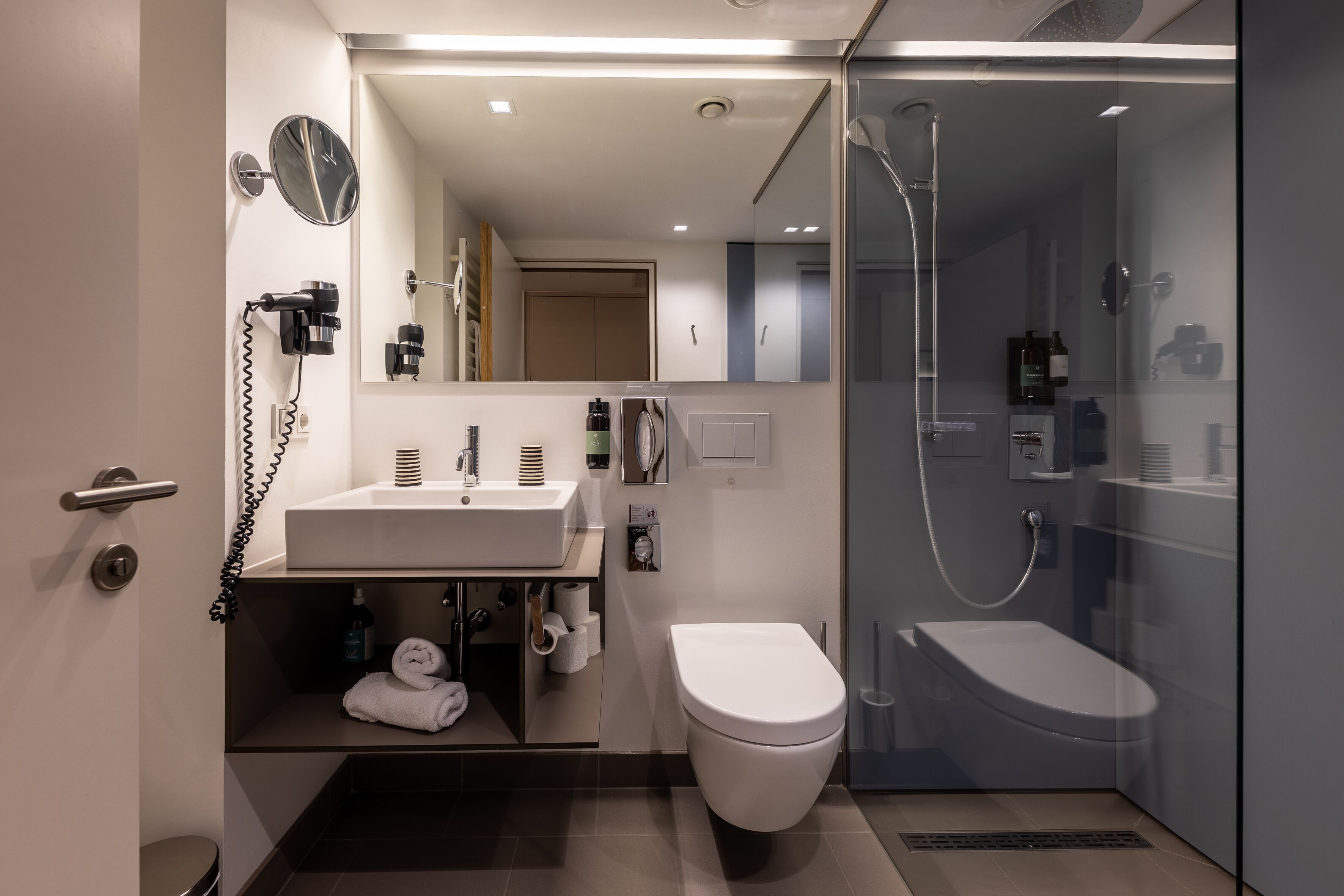Un baño moderno con ducha de vidrio, inodoro blanco y tocador con espejo y secador de pelo.