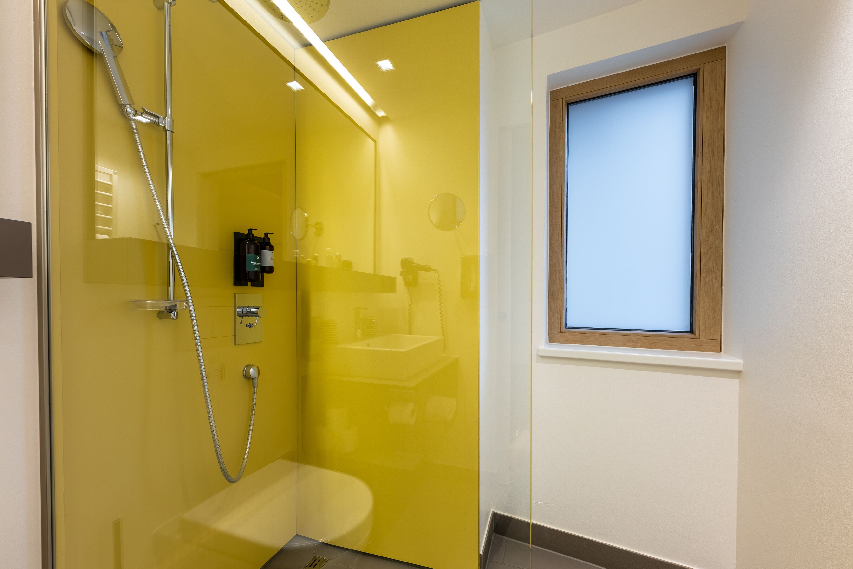 A bathroom with a yellow wall, a shower, a sink, and a window.