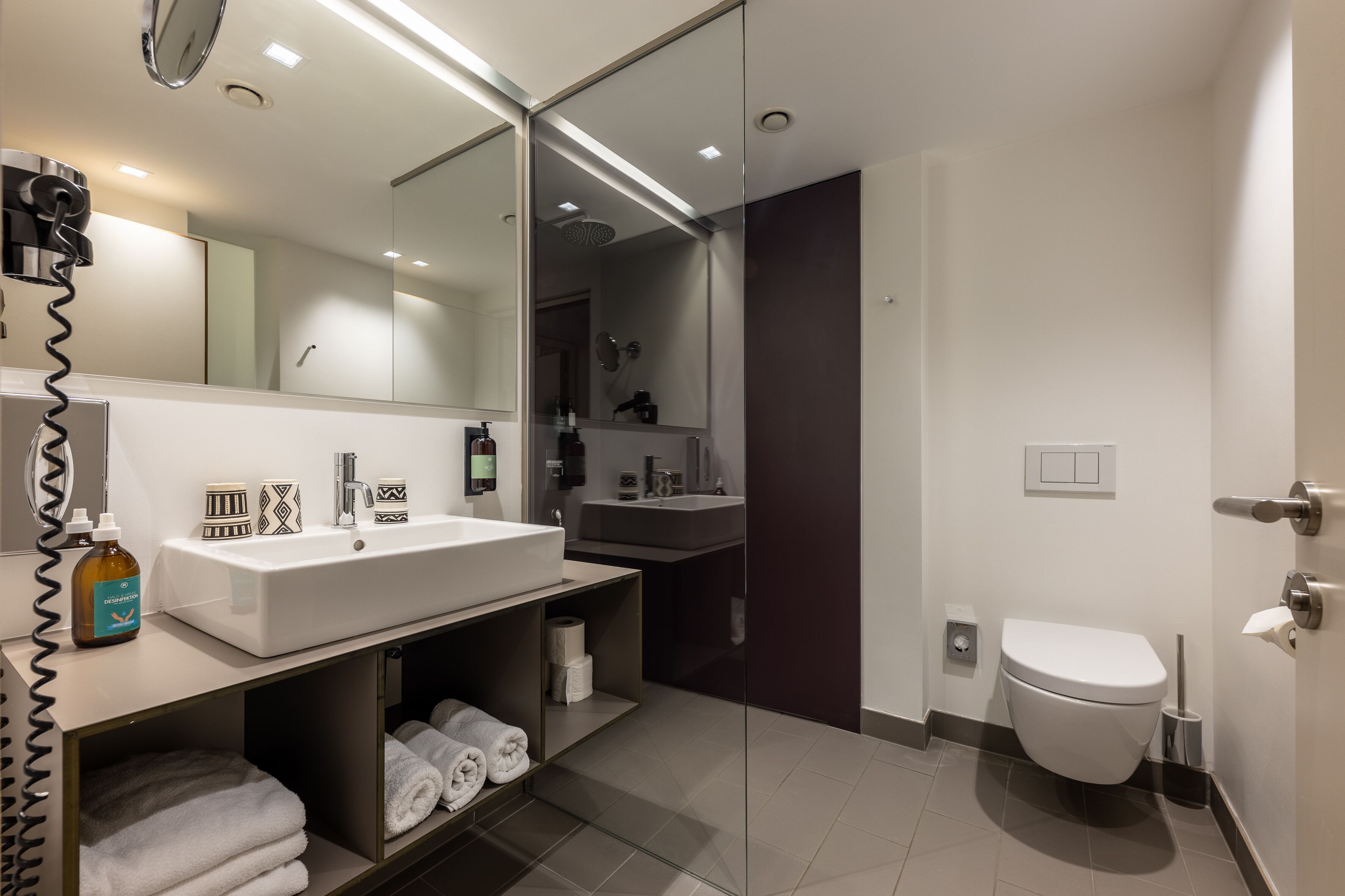 Une salle de bain moderne avec un grand miroir, un lavabo blanc et des toilettes. On y trouve des serviettes et des articles de toilette sur le comptoir.