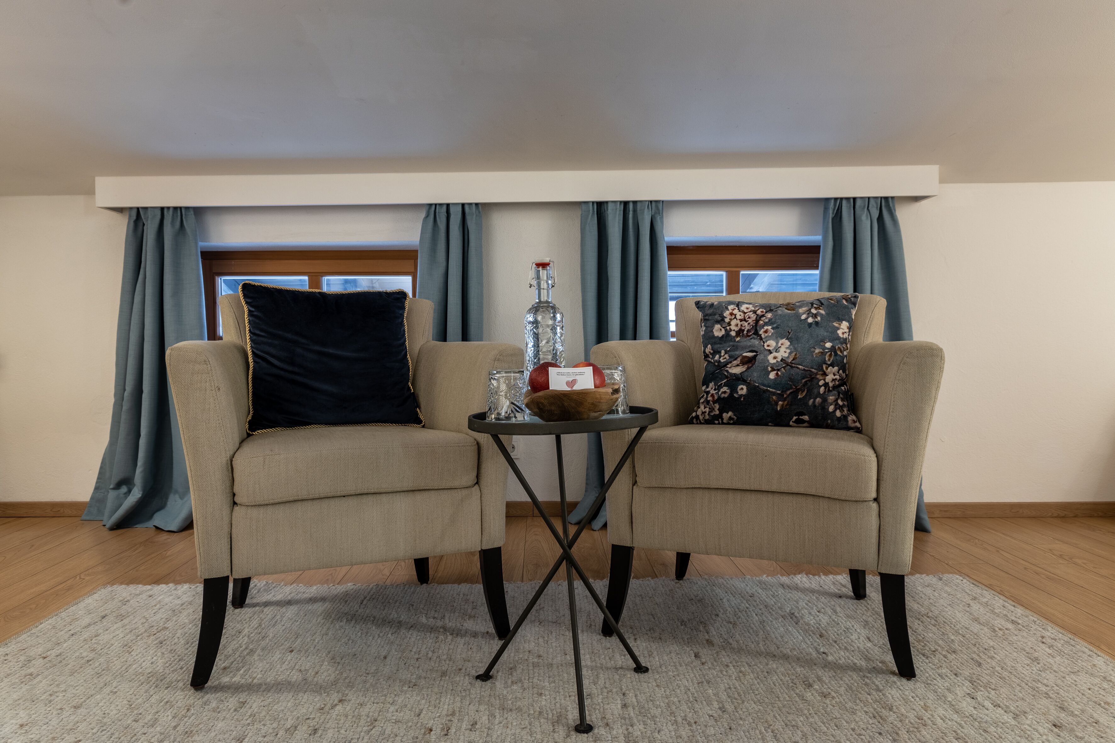Une pièce avec deux fauteuils beiges, une petite table sur laquelle se trouvent une bouteille, des verres et un bol. Il y a des rideaux bleus, une fenêtre et un plancher en bois.