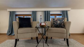 Een kamer met twee beige fauteuils, een tafeltje met een fles, glaasjes en een schaal. Er zijn blauwe gordijnen, een raam en een houten vloer.