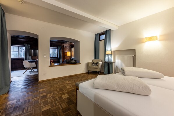 A hotel room with a wooden parquet floor, a bed with white linens, a chair, a desk with a lamp, and a view of the outside through a window. - Hotel & Wirtshaus Post (Sankt Johann in Tirol)