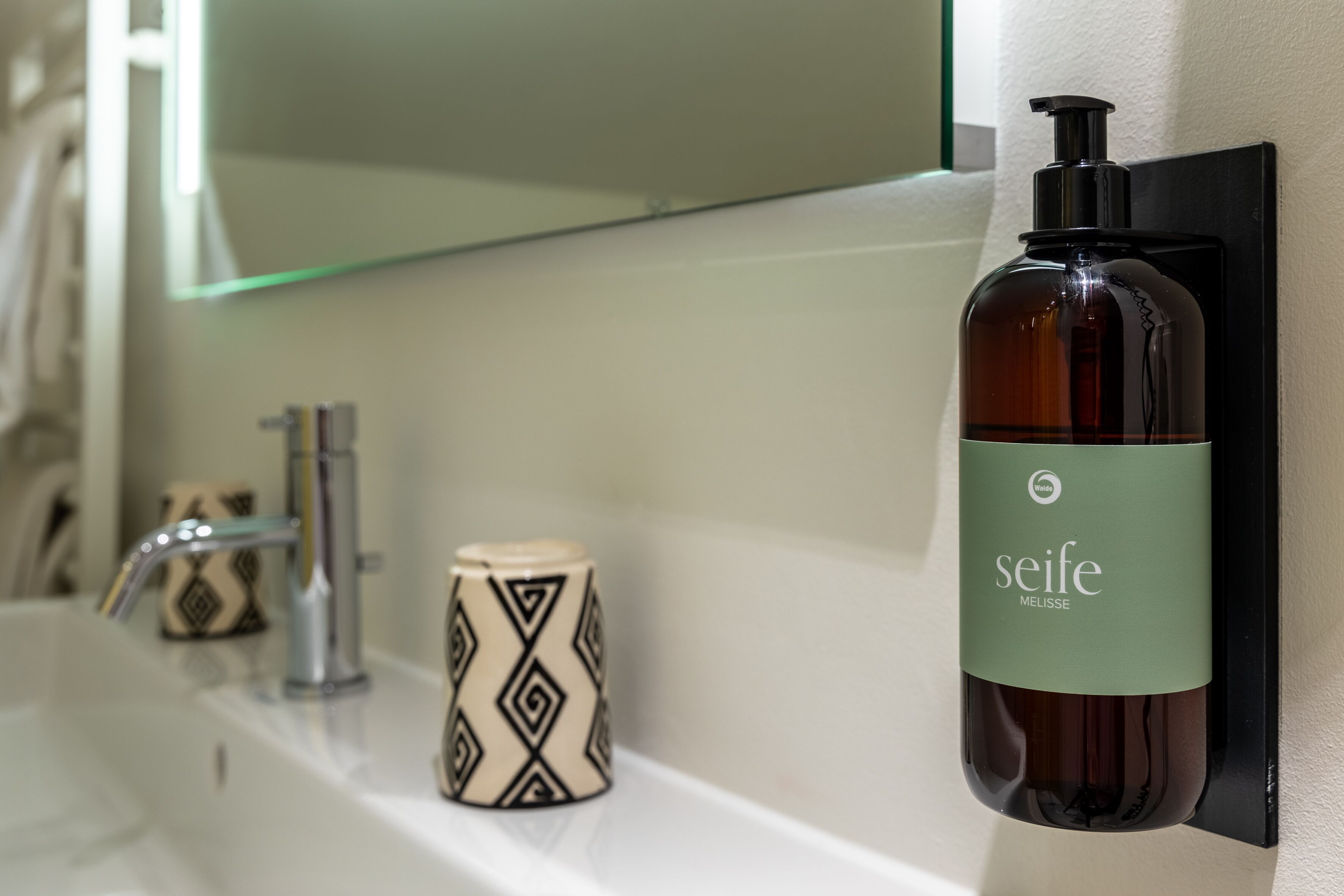 Une salle de bain avec un savon Seife Melissa dans un distributeur mural, une tasse à motifs et un robinet moderne.