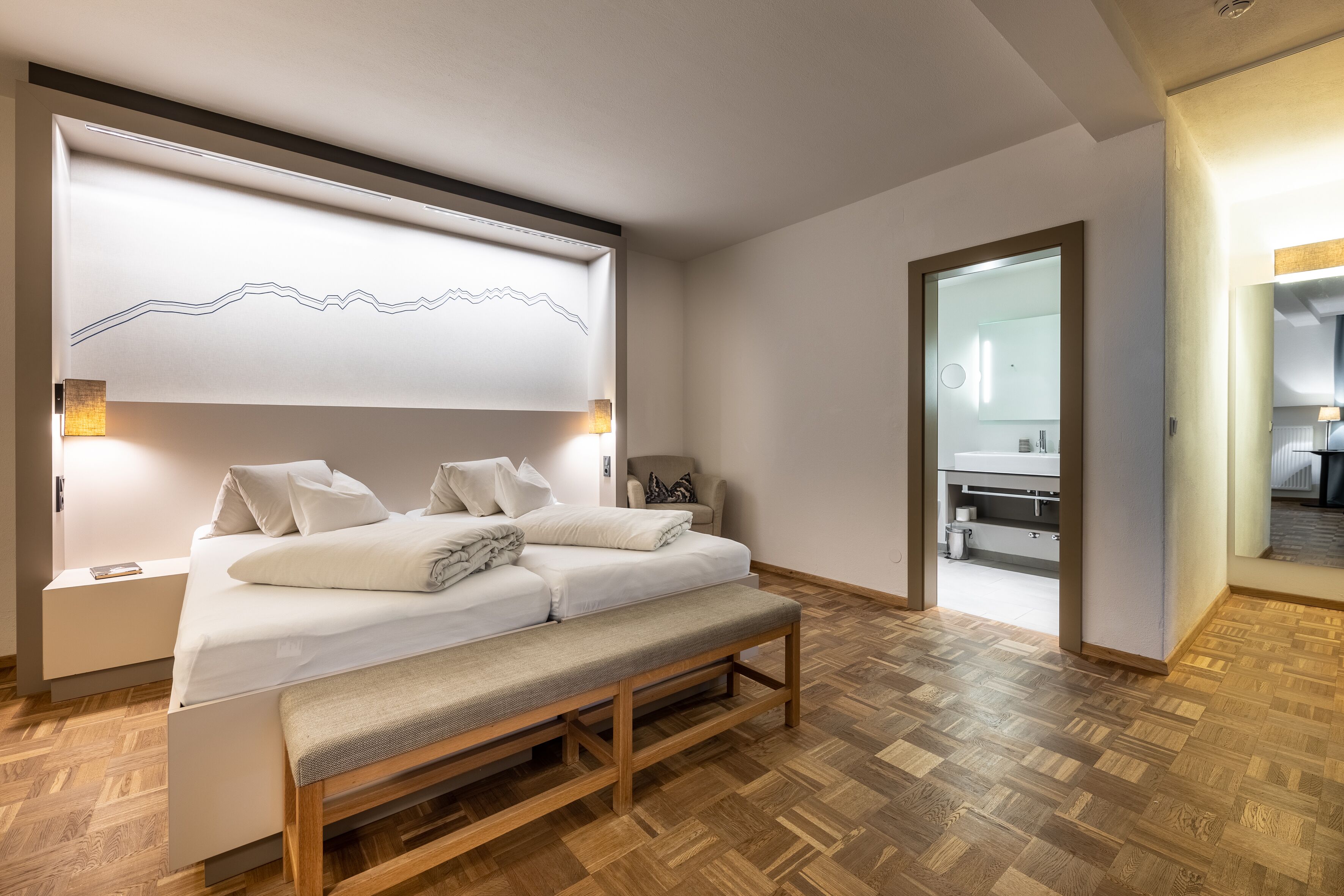 Un dormitorio moderno con cama, banca, mesitas de noche y una lámpara fijada a la pared.