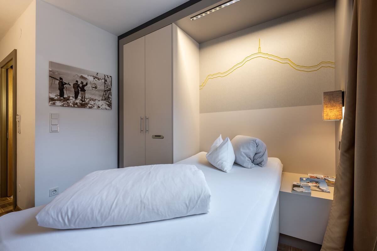 A hotel room with a bed, a nightstand with books, a wardrobe, and a wall-mounted light fixture.