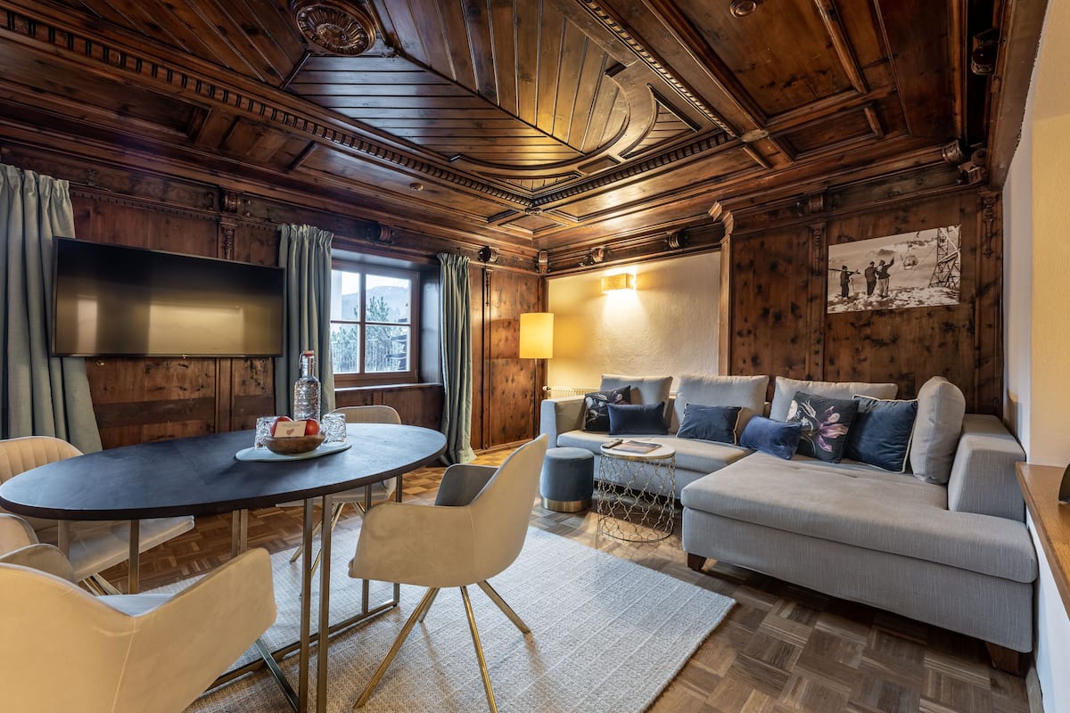 A cozy room with a wooden ceiling, a round dining table with chairs, a sofa, and a TV mounted on the wall.