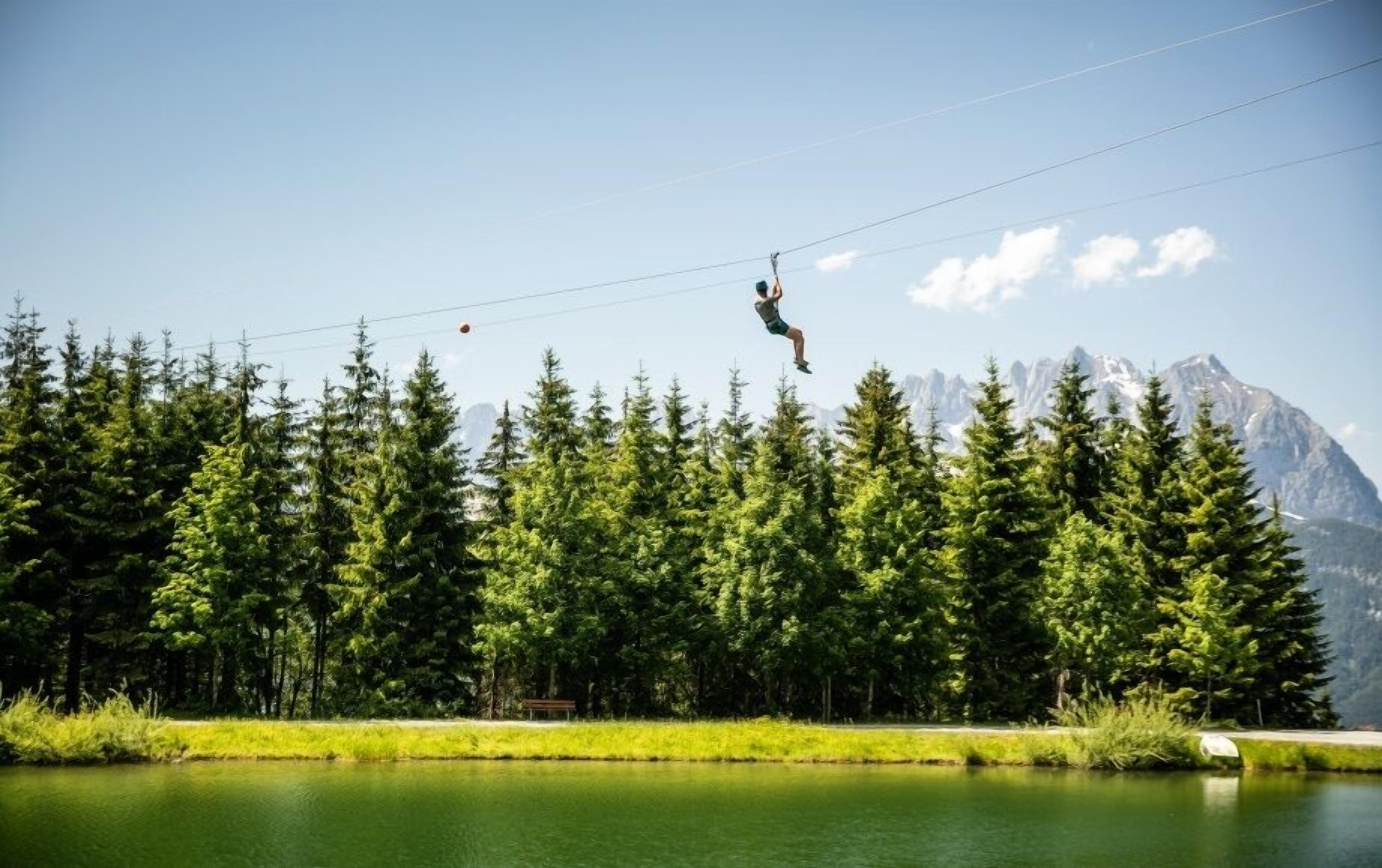 A person zip-lining over a forest with a lake and mountains in the background.