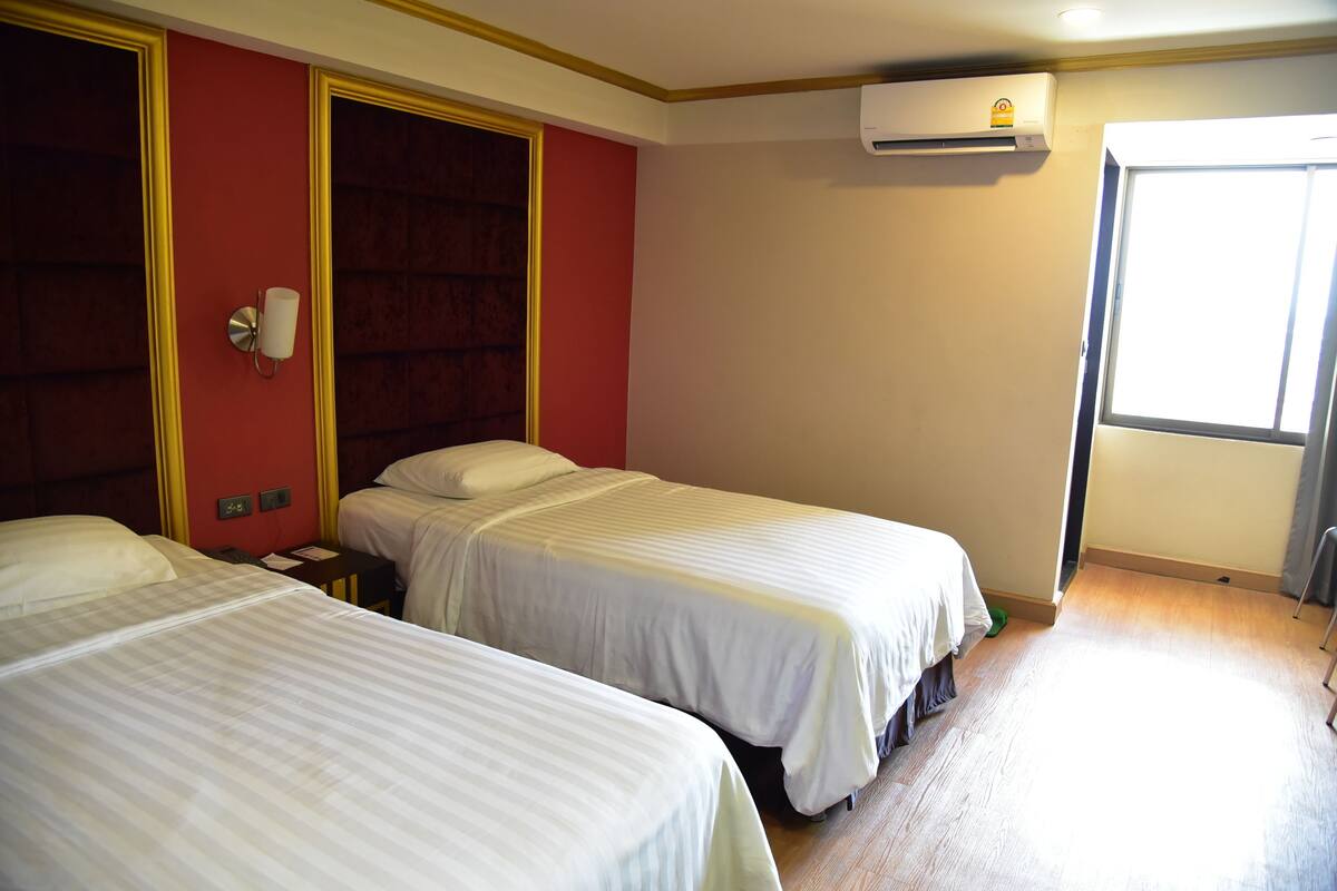 A hotel room with two beds, a red accent wall, an air conditioning unit, and a window.