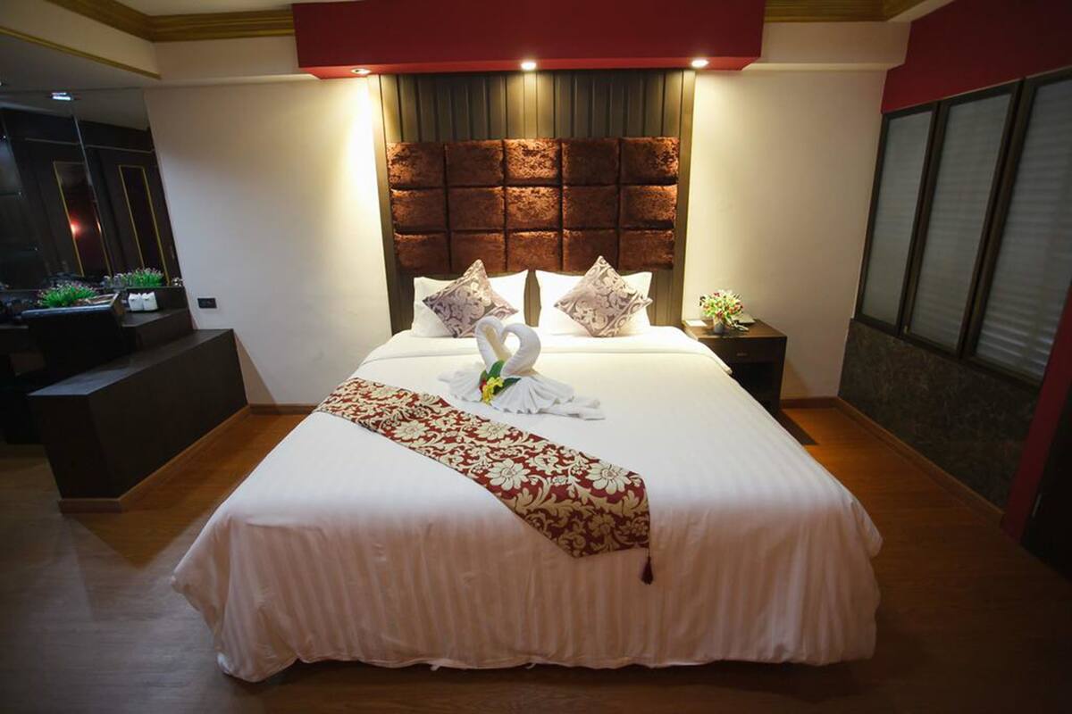 A neatly made bed with a decorative swan pillow arrangement, a headboard with a textured pattern, and a bedside table with a vase of flowers.