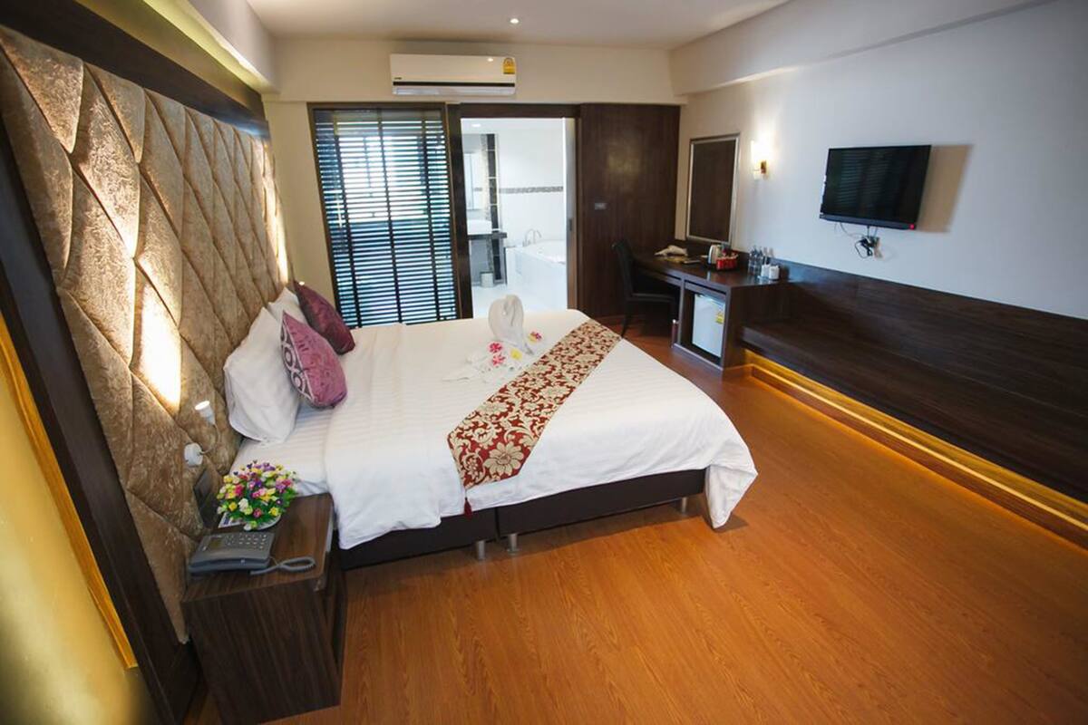 A hotel room with a large bed, a desk with a chair, a TV, and a bathroom visible through a sliding door.