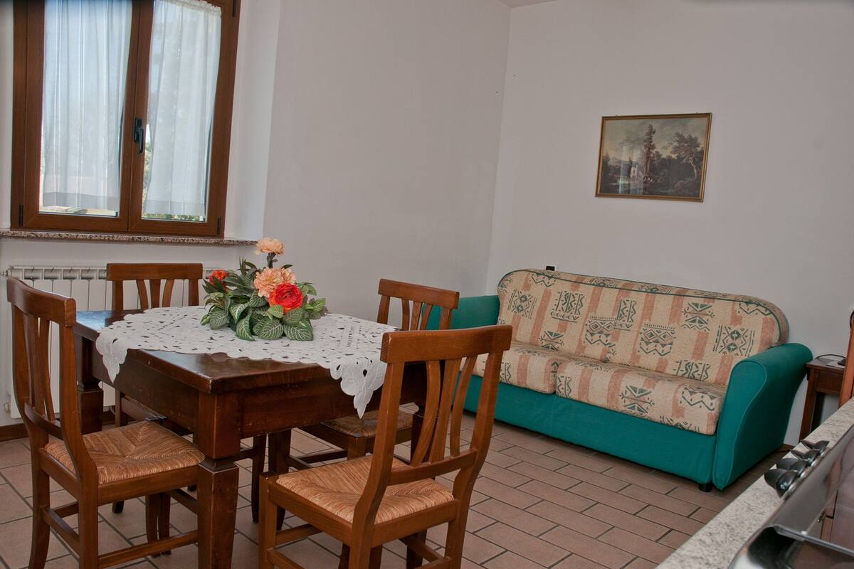 Un coin repas avec une table en bois, une nappe en dentelle et des chaises. On y trouve également un canapé avec un coussin à motifs et un tableau accroché au mur.
