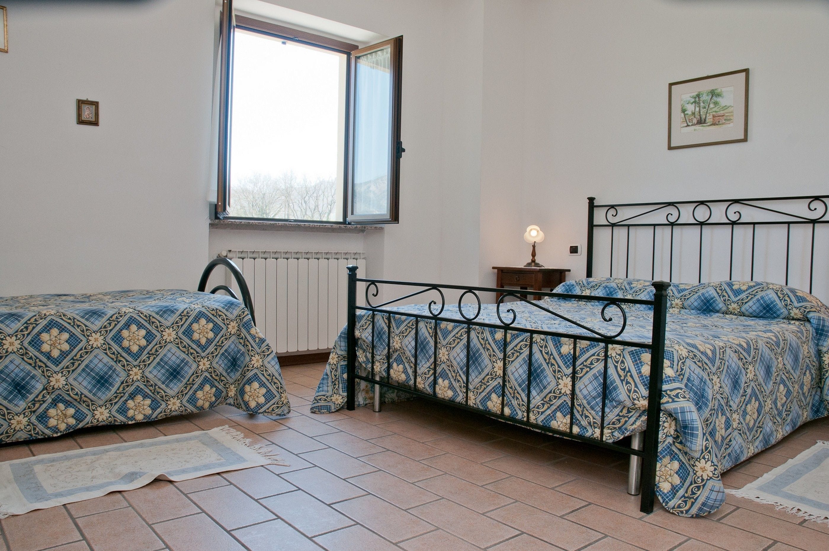 A room with two beds, a window, a radiator, and a small table with a lamp.