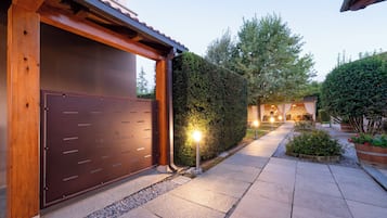 Una entrada protegida con estructura de madera, un camino que lleva a una zona iluminada y un seto bien cuidado.