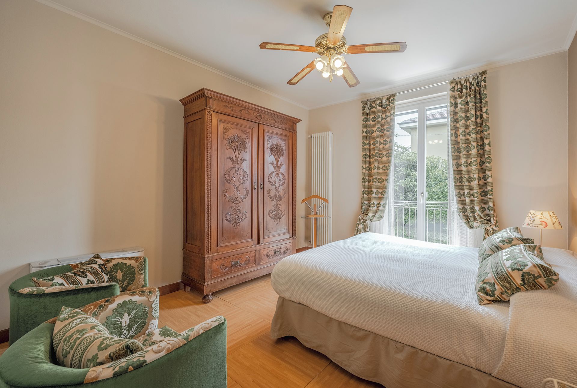 A bedroom with a bed, a wooden wardrobe, a green armchair, and a ceiling fan.
