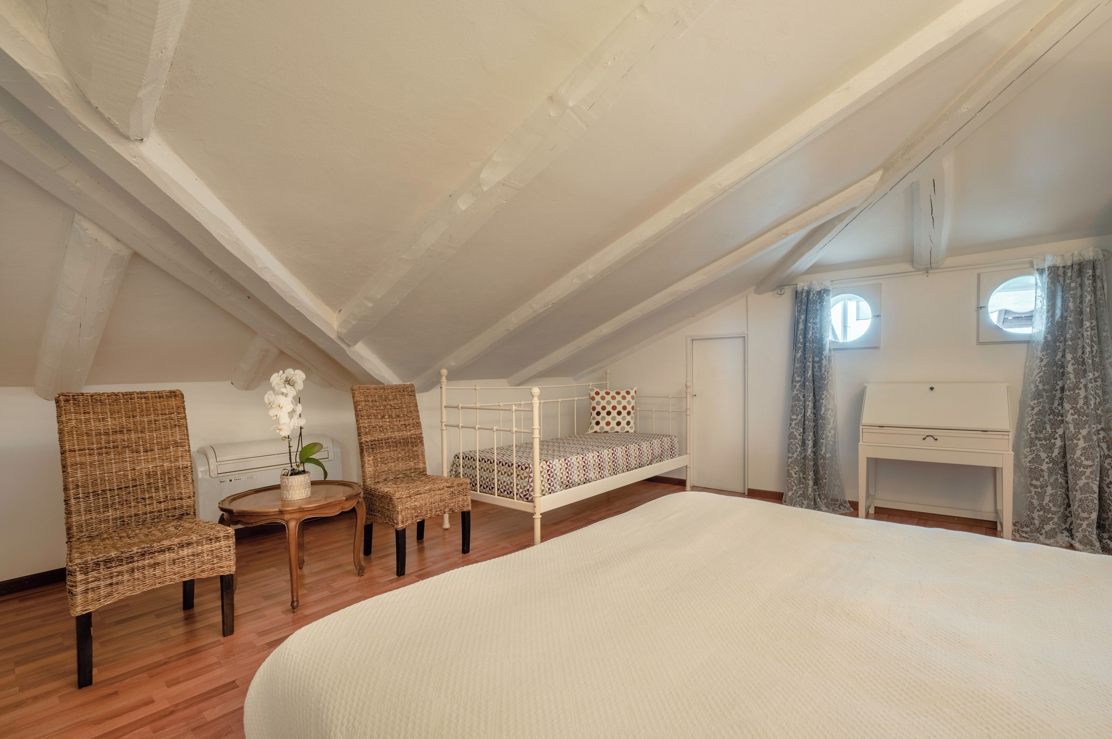A bedroom with a bunk bed, two wicker chairs, a small table with a plant, a bedside table, and a window with curtains.