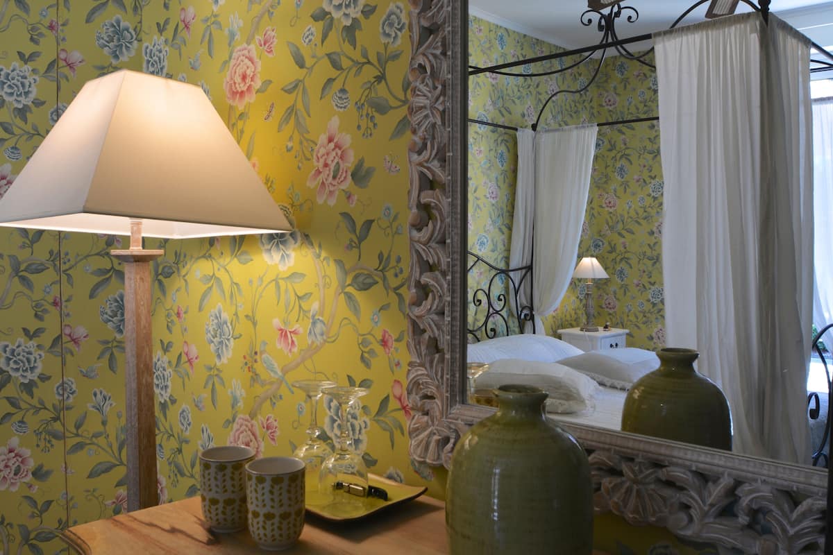 Una habitación con papel tapiz floral, una mesita de noche con lámpara, un espejo y una cama con dosel.