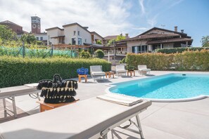 A swimming pool area with lounge chairs and a bench, surrounded by well-maintained hedges and a building with a clock tower in the background. - La Rosa Gialla (Barolo)