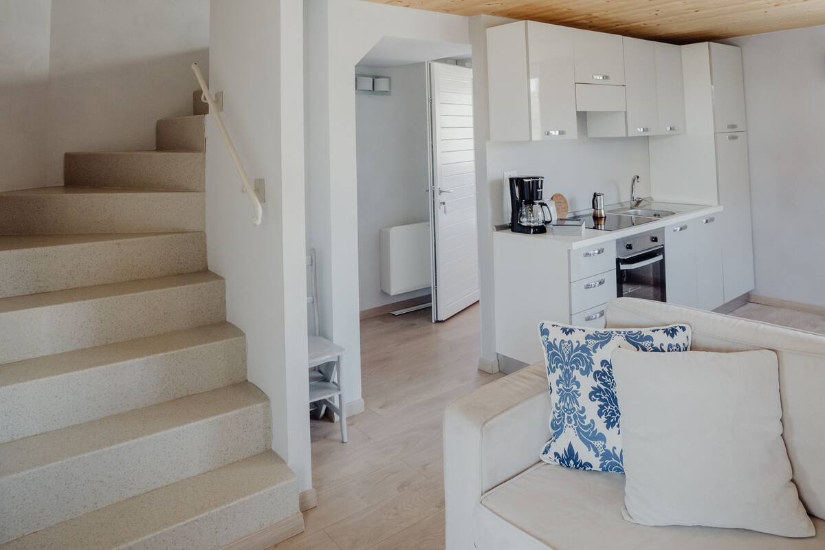 Un interior moderno con una escalera, una zona de cocina y un espacio de estar con un sofá y cojines estampados.