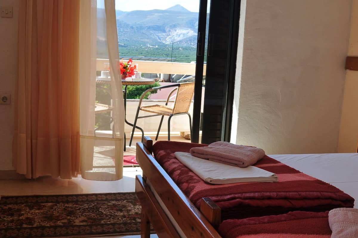 A bedroom with a bed, a window with curtains, a view of the outdoors, and a small table with chairs.