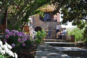 A stone building with a balcony, surrounded by lush greenery and flowering plants. - House Margot (Sitia)
