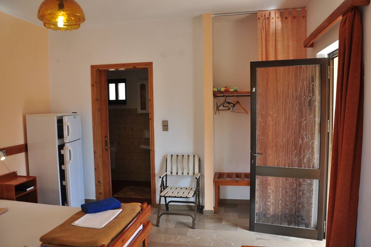 A compact room with a bed, a small table, a chair, a wooden bench, a hanging chair, a wardrobe, and a door with a bamboo-patterned glass panel.