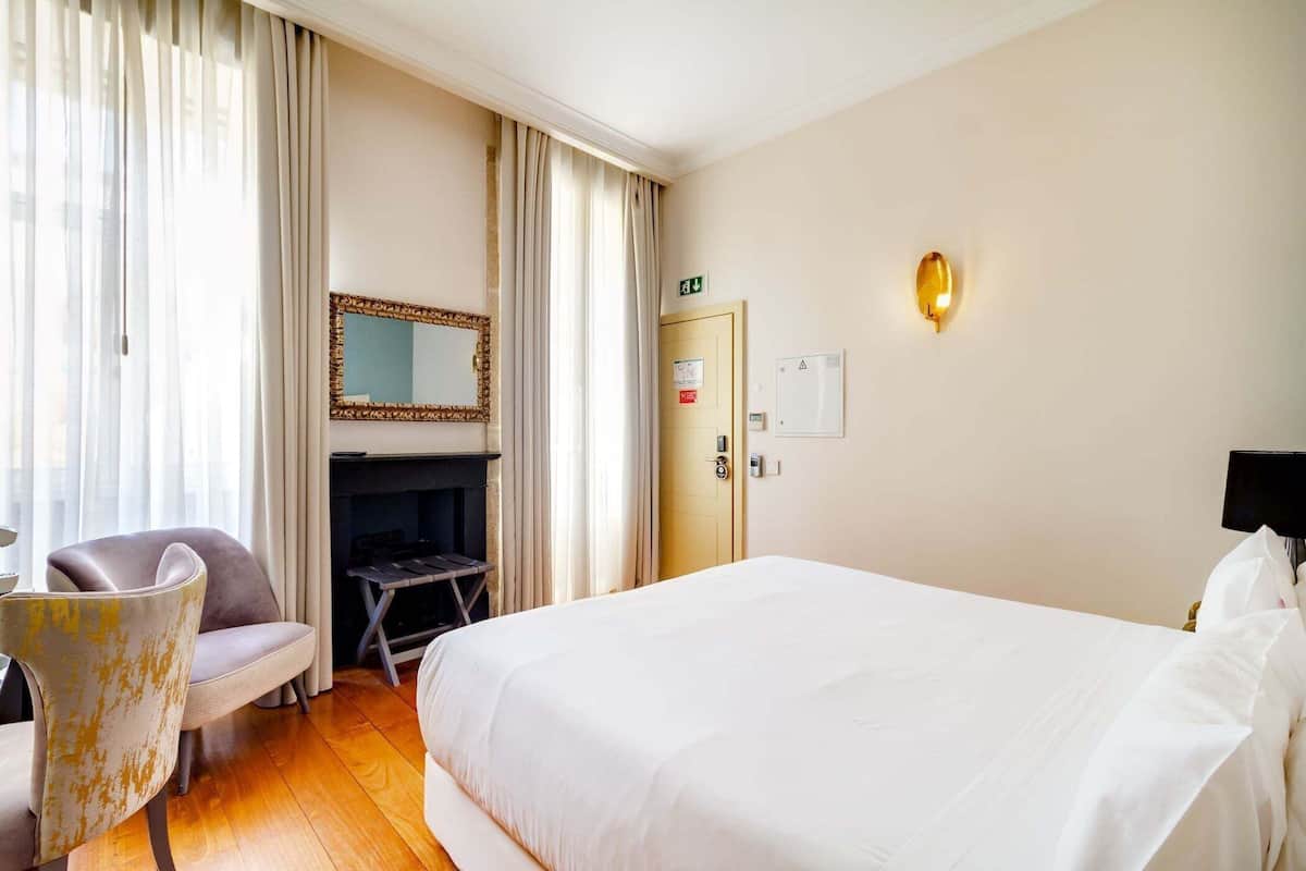 A hotel room with a bed, a chair, a fireplace, a mirror, a door, and a wall-mounted light fixture.