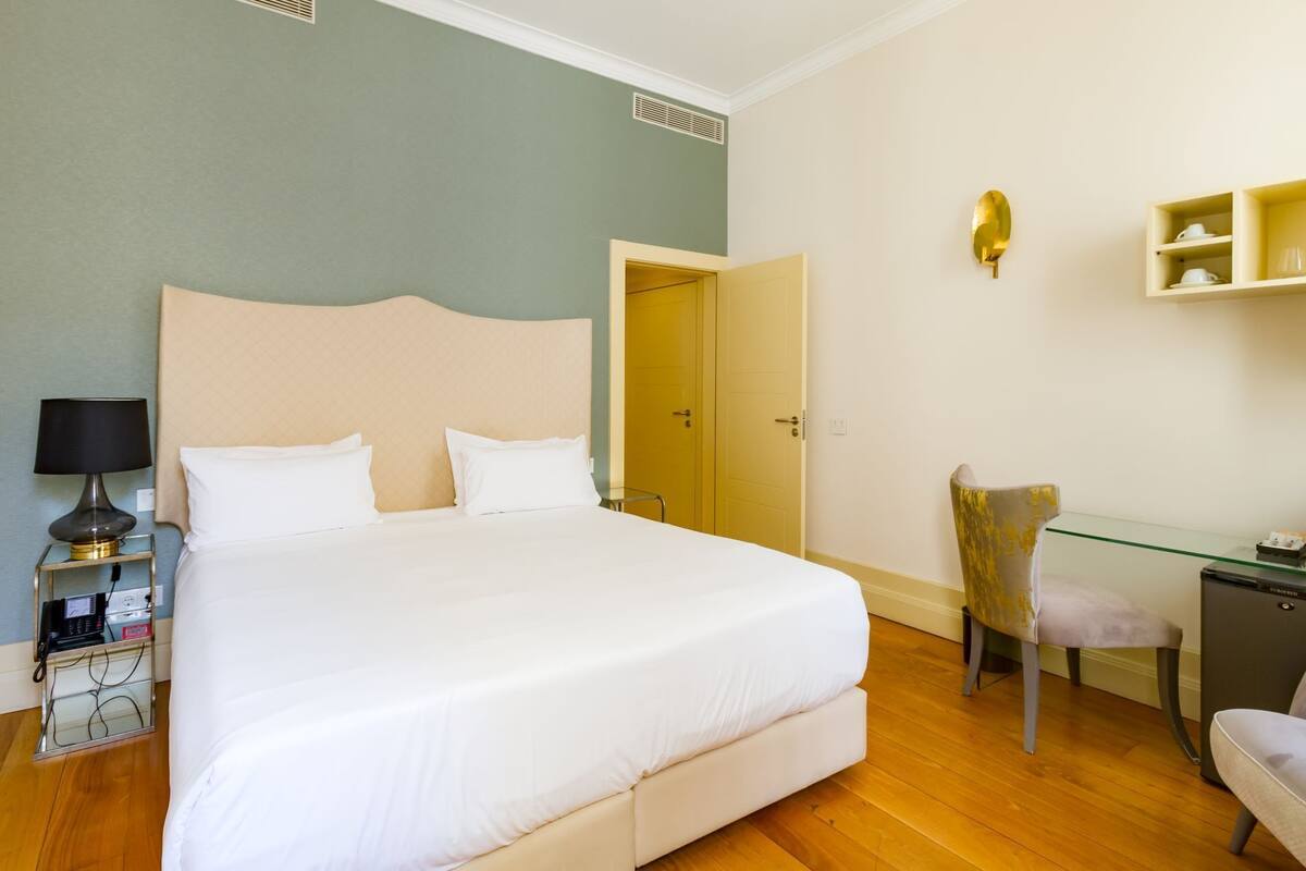 A neatly arranged hotel room with a large bed, bedside table with a lamp, a desk with a chair, and a small shelf with cups.