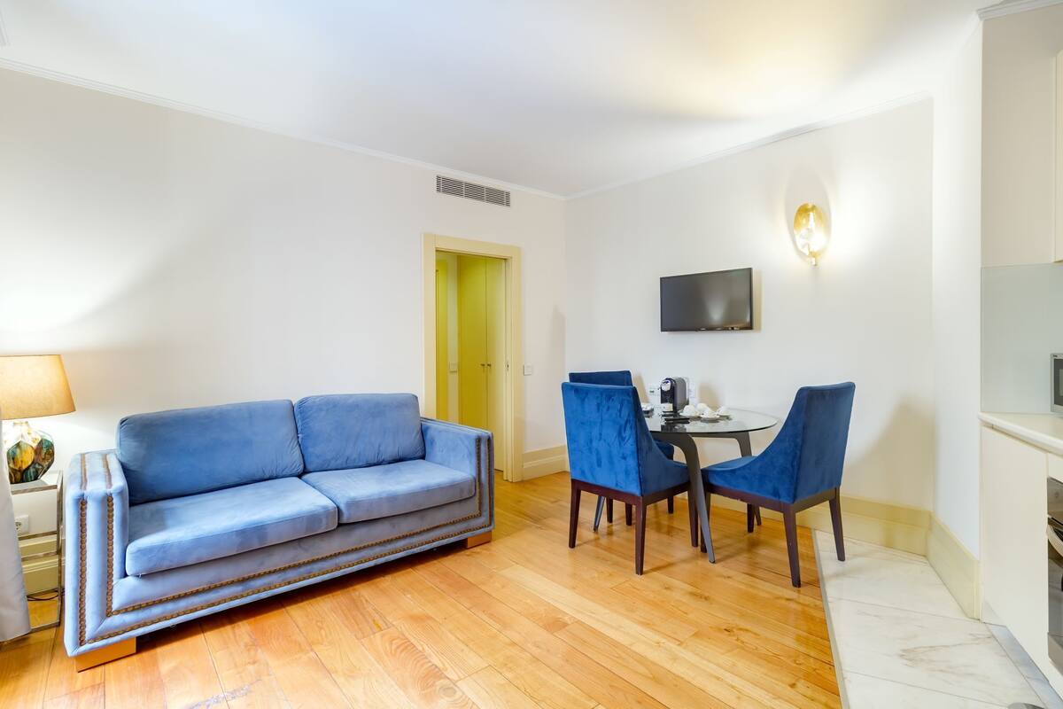 A living room with a blue sofa, two blue chairs, a small round table, a TV mounted on the wall, and a kitchen area with a stove.