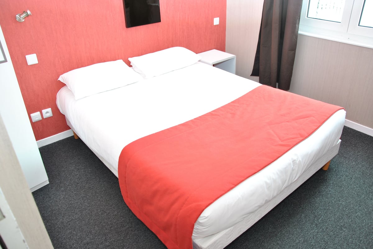 Un lit bien fait, avec une literie blanche et rouge, une table de chevet et un luminaire fixé au mur.