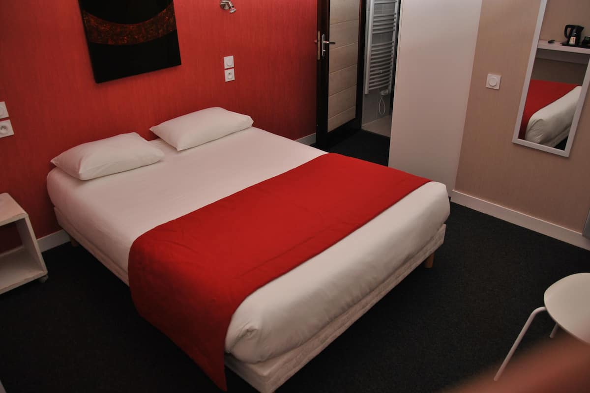 Une chambre d’hôtel avec un lit, une couvre-lit rouge et blanche, une table de chevet blanche, un mur rouge, une porte, un miroir et une petite table blanche.