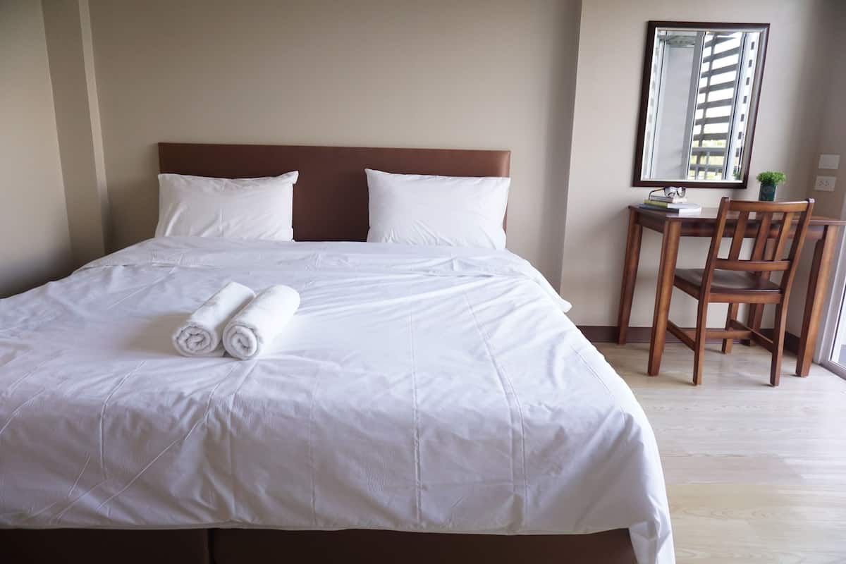 A neatly made bed with white linens and rolled towels, a wooden headboard, a small desk with a chair, and a mirror on the wall.