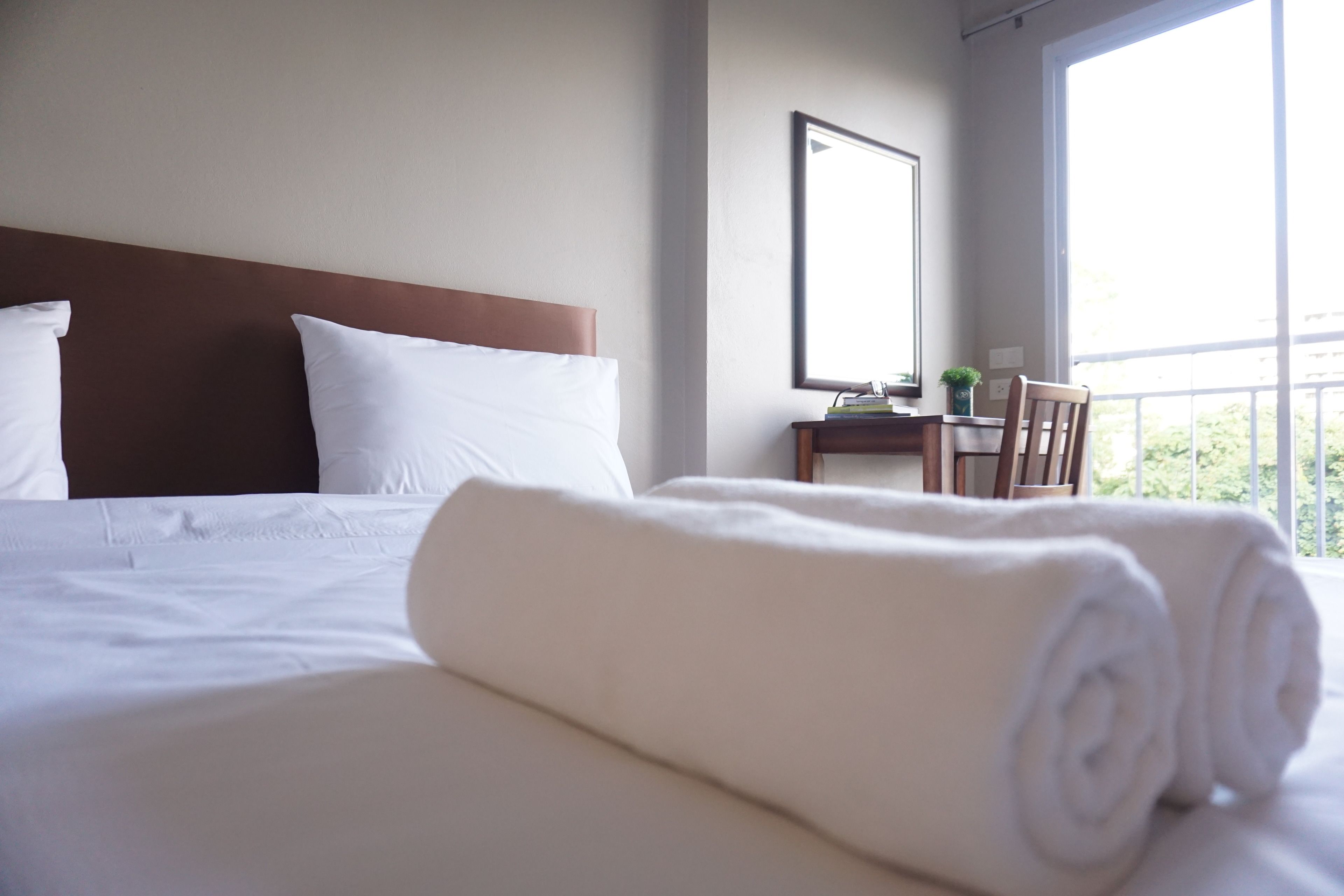 Una cama bien hecha con sábanas blancas y toallas enrolladas, un cabecero de madera, una mesita con una planta y una ventana con vistas al verdor.