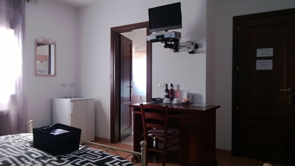 Una habitación pequeña y sencilla con cama, un escritorio y una silla, un televisor montado en la pared, un refrigerador y una puerta con un cartel.