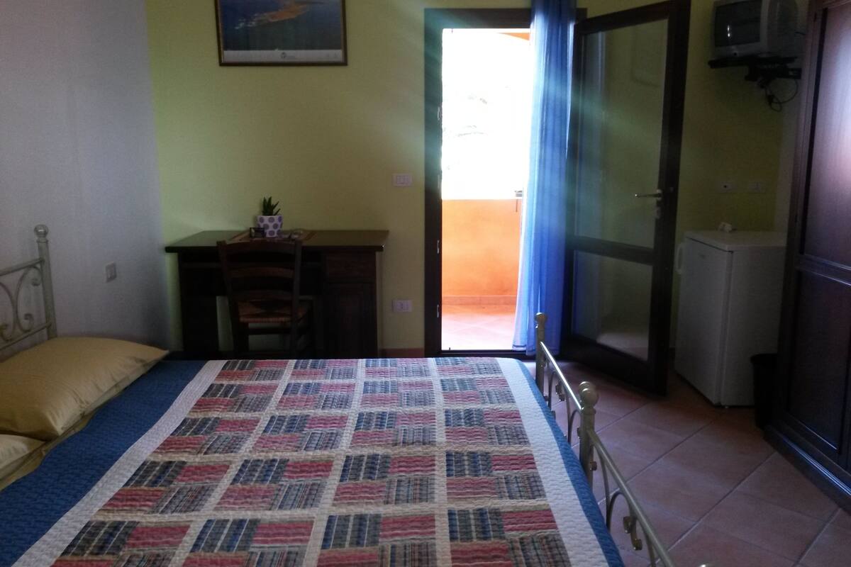 A bedroom with a bed, a desk, a chair, a television, and a door leading to an outdoor area.