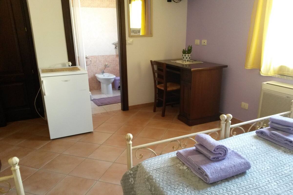 A bedroom with a bed, a desk, a chair, a small white refrigerator, and a bathroom visible through an open door.