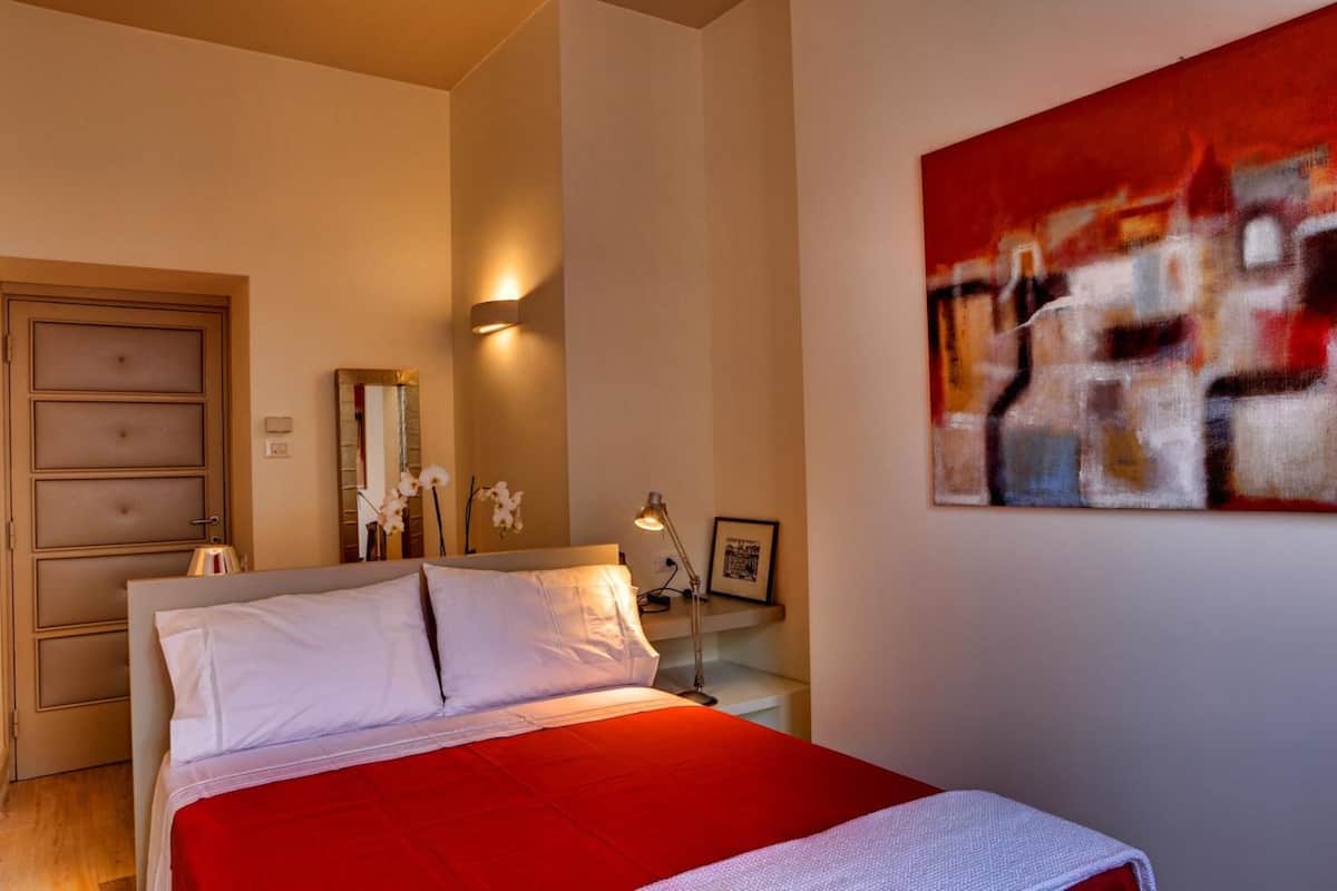 A bedroom with a bed, a nightstand, a wall-mounted lamp, a painting, and a door.