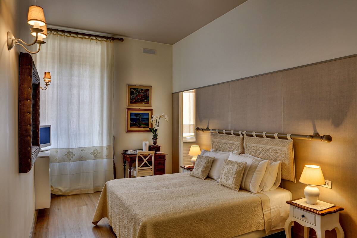 A bedroom with a large bed, bedside lamps, a desk with a computer, and a window with curtains.