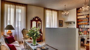 Una sala de estar con un sofá, una mesa con libros, un florero con flores, una lámpara, un espejo y una estantería.