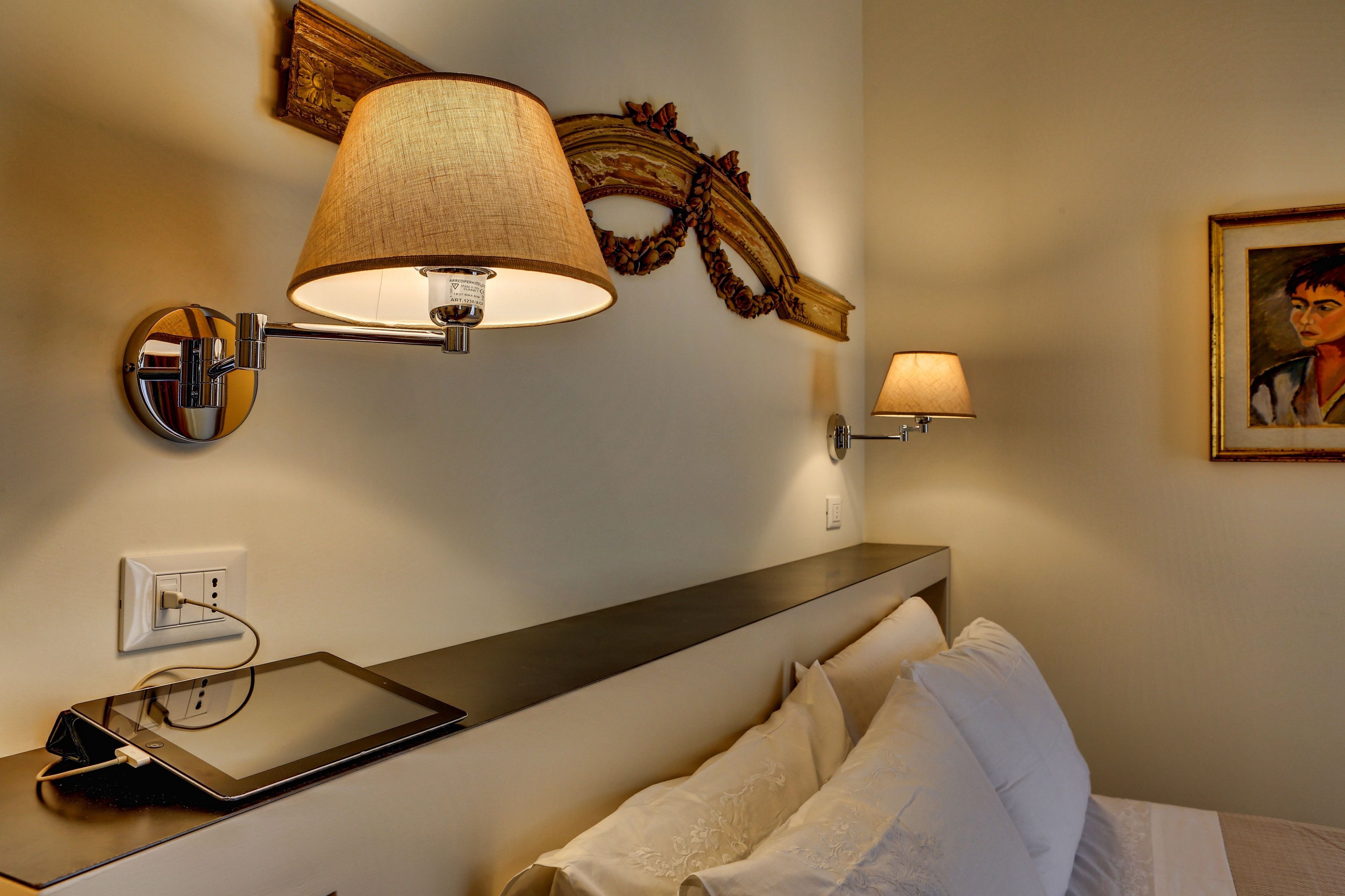 Hotellihuone, jossa on yöpöytä, lamppu, taulu ja seinälle kiinnitetty peili.