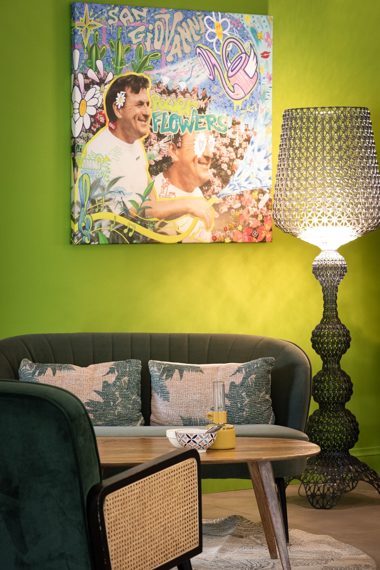 Een woonkamer met een groene muur, een bank met gestreepte kussens, een tafel met een schaal en een lamp, en een kleurrijk schilderij boven de bank.