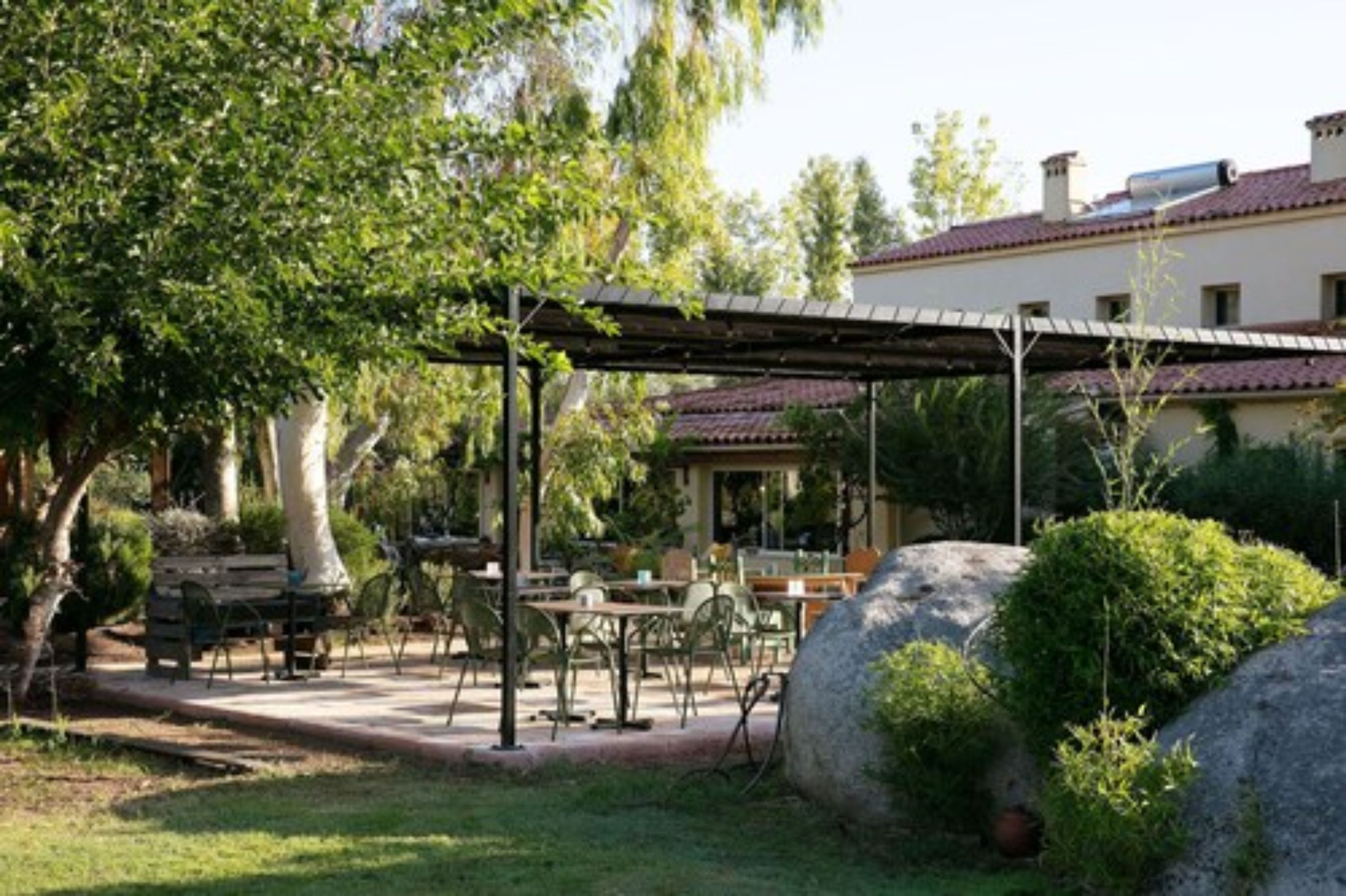 Un'area patio esterna con tavoli e sedie, circondata dal verde e da un edificio con tetto in tegole rosse.